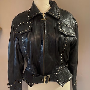 May include: Black leather jacket with gold stud detailing. The jacket has a notched collar, a zip-up front, and a belted waist. It features two front pockets and decorative stitching.