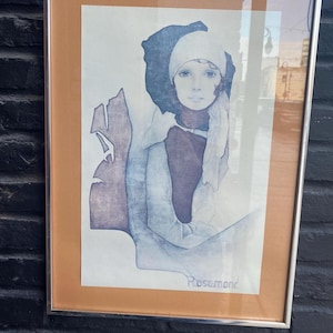 Puede incluir: Obra de arte enmarcada que presenta un retrato de una persona con un gorro y bufanda blancos. La obra está en tonos azules, morados y marrones, con el nombre "Rosemond". El marco es plateado.