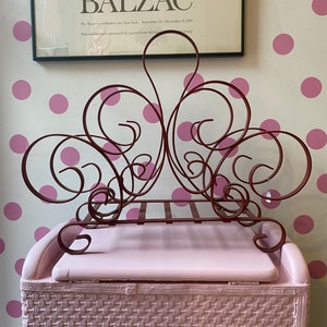 May include: A red metal magazine rack with a decorative scroll design sits on top of a pink wicker storage bin. The rack has four shelves and is in the shape of a heart. The bin has a woven pattern and is in the shape of a rectangle.