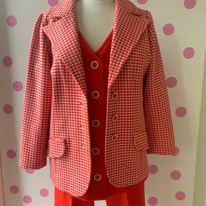 Puede incluir: Una chaqueta de color rojo y blanco con estampado de pata de gallo con una falda y pantalones a juego en rojo. La chaqueta tiene un cuello de muesca, mangas largas y cierre de botón. La falda y los pantalones son de color rojo sólido.