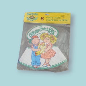 Peut inclure: Un paquet de six chapeaux de fête Cabbage Patch Kids. L'emballage présente des illustrations de deux personnages Cabbage Patch Kids, un garçon en salopette et une fille en robe rose. Les chapeaux sont coniques avec un motif à rayures.