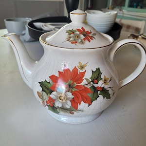 May include: A white ceramic teapot with a lid, decorated with a floral pattern of red poinsettias, green holly leaves, and gold trim. The teapot has a curved spout and handle, ideal for serving tea.