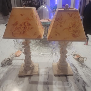Puede incluir: Par de lámparas de mesa de color marfil. Cada lámpara presenta una pantalla cuadrada con un fondo amarillo claro y un diseño de querubines. La base está intrincadamente esculpida con figuras y descansa sobre una base cuadrada de madera. Se ven cables eléctricos.