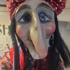 May include: A doll with a long, pointed nose, pale skin, and rosy cheeks. The doll has dark hair, blue eyes, and wears a red head covering with white polka dots. The doll is dressed in a patterned garment with red, blue, and orange colours.
