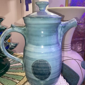 May include: A tall, ceramic jug with a lid, handle, and spout. The jug is a marbled blue and white with a dark blue planet design on the front. The handle has a unique, decorative shape.
