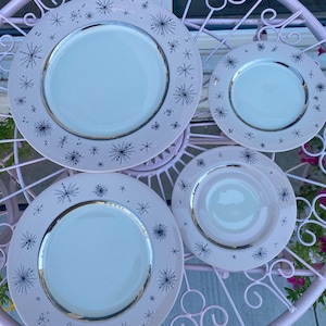 Peut inclure: Quatre assiettes en céramique rose clair avec un centre blanc et un bord argenté. Les assiettes sont décorées d'un motif d'éclats d'étoiles bleu foncé et de petits points. Les assiettes sont disposées sur une table en métal rose.