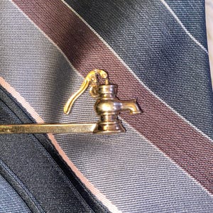 May include: A gold-toned tie clip featuring a miniature water pump design. The clip is set against a striped tie in shades of navy, gray, and burgundy, creating a classic and elegant accessory.