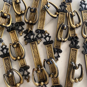 Puede incluir: Una colección de tiradores de armario vintage. Cada tirador presenta una placa trasera rectangular texturizada en tono dorado con un acento negro decorativo en la parte superior e inferior. Los tiradores tienen un asa arqueada en tono dorado.