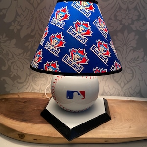 May include: A baseball-themed lamp with a white baseball base, red stitching, and a blue and red MLB logo. The blue lampshade is printed with the Toronto Blue Jays logo. The lamp is set on a black and white home plate base.