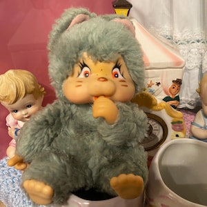 May include: A vintage plush toy with a light blue furry body, large eyes, and a small orange nose and mouth. The toy is sitting in a decorative ceramic container. Other vintage figurines and a circus-themed cookie jar are in the background.