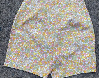 May include: A pair of white shorts with a floral print in shades of pink, yellow, and green. The shorts have a white waistband and a white zip.