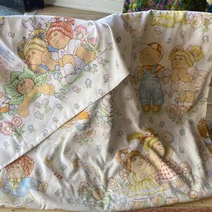 May include: A white fabric sheet featuring a repeating pattern of Cabbage Patch Kids characters, including children with yarn-like hair, wearing overalls and dresses, surrounded by colorful flowers, butterflies, and a kite, with the logo text visible.