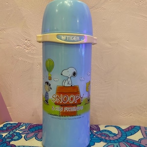 Puede incluir: Una botella de agua aislada azul claro con Snoopy y sus amigos. La botella tiene una tapa azul claro y una banda color crema con la palabra "TIGER". La botella tiene una ilustración de dibujos animados de Snoopy y otros personajes. También es visible el texto "SNOOPY & HIS FRIENDS".