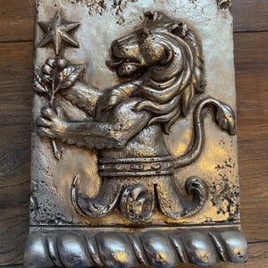 Könnte beinhalten: Silberfarbene rechteckige Reliefskulptur mit einem heraldischen Löwen, der einen Stern und einen Zweig hält. Der Löwe hat eine detaillierte Mähne und trägt einen dekorativen Gürtel. Der Sockel hat ein seilartiges Design.