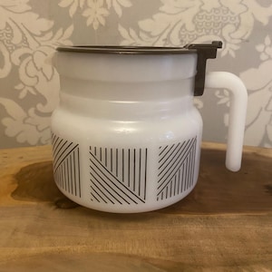 Op de afbeelding: Witte emaille koffiepot met een zwart deksel en handvat. De pot heeft een zwart geometrisch ontwerp op de onderste helft. De pot staat op een houten oppervlak.