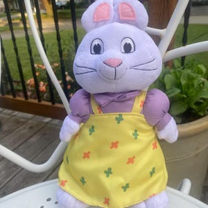 May include: A plush rabbit toy with light purple fur, pink inner ears, and a pink nose. The rabbit wears a yellow jumper dress featuring a pattern of small green and orange cross-shaped icons, layered over a light purple short-sleeved shirt.