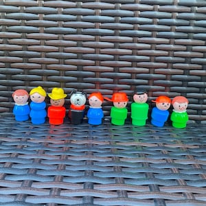 Könnte beinhalten: Eine Sammlung von Vintage Fisher-Price Little People Figuren. Die Figuren haben verschiedene farbige Outfits und Hüte, darunter Blau, Rot, Gelb und Grün. Eine Figur ist ein Hund. Die Figuren sind in einer Reihe angeordnet.