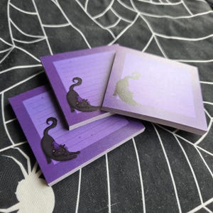 May include: Square purple notepads with a gradient design and a black cat illustration wearing a witch hat. The notepads have lined pages and are stacked on a black and white spiderweb patterned surface.