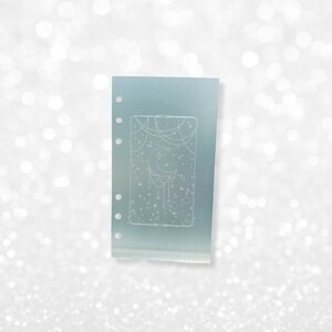 May include: A light blue planner page with a white design of a crescent moon with stars and a string of beads hanging from it.