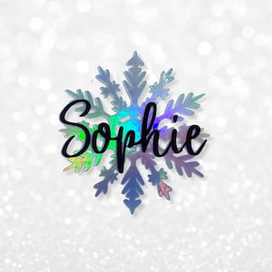 May include: An iridescent snowflake ornament with the name "Sophie" in black script. The snowflake has a detailed, multi-pointed design, with a shimmering, colour-shifting effect. The background is a soft, white, blurred pattern.