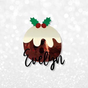 Pode incluir: Um enfeite de pudim de Natal com uma superfície brilhante e reflexiva. O enfeite apresenta uma cobertura de creme branco, bagas vermelhas e folhas verdes de azevinho. O nome "Evelyn" está escrito em itálico na parte inferior.