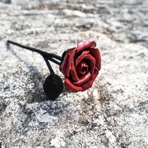 Può includere: Una singola rosa di metallo rosso con uno stelo e foglie di metallo nero. La rosa è sdraiata su una superficie grigia testurizzata.