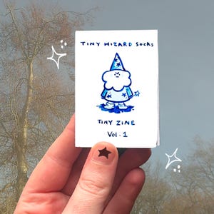 May include: A small, white zine titled "Tiny Wizard Socks" featuring a blue cartoon wizard with a star-topped hat and a wand. The zine is held in a hand with a star-shaped nail sticker. The text "Tiny Zine Vol. 1" is also visible.