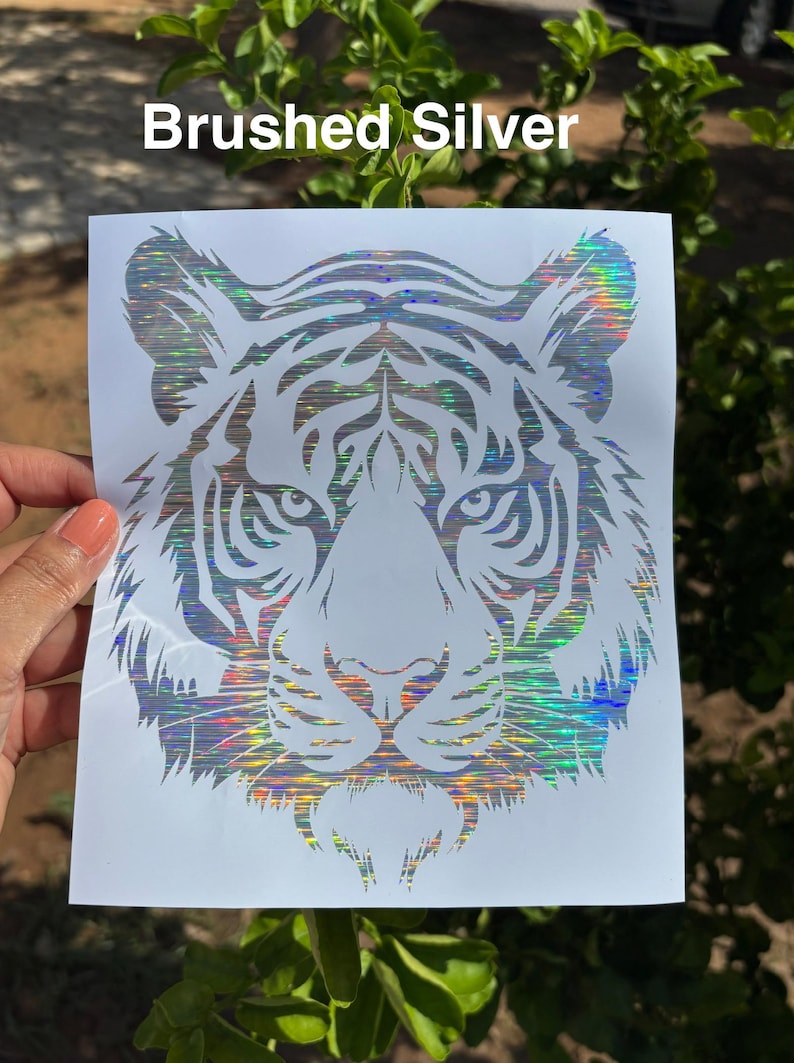 May include: A decal featuring a stylized tiger head design with a holographic, brushed silver finish. The tiger graphic is printed on a white rectangular backing sheet. The text "Brushed Silver" is visible at the top of the image.