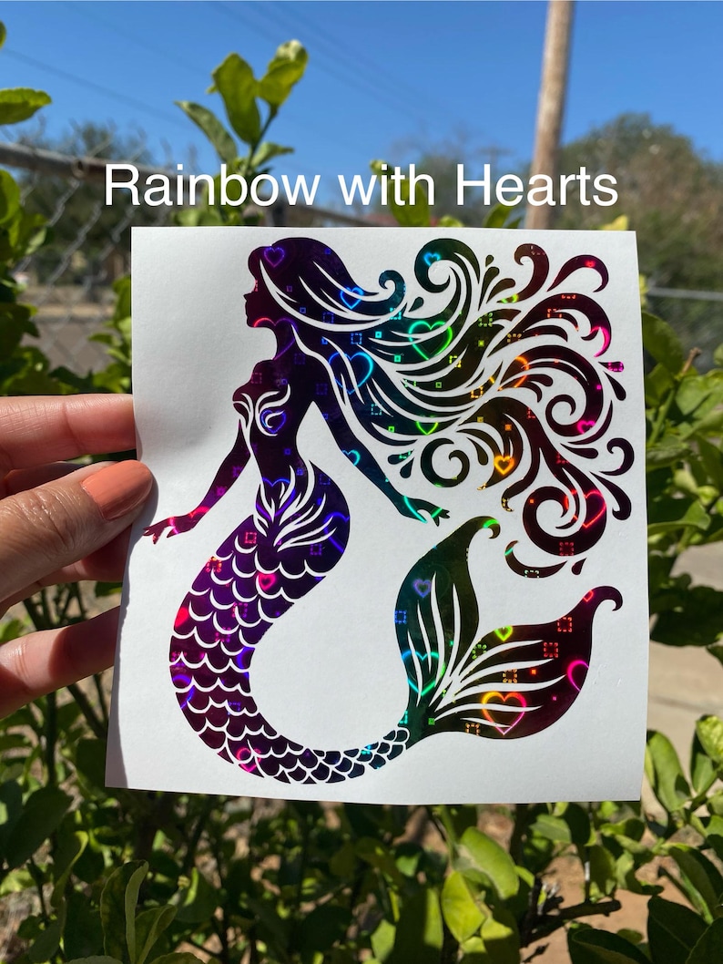 K&ouml;nnte beinhalten: Ein holografischer Meerjungfrau-Aufkleber mit dem Text "Rainbow with Hearts". Die Meerjungfrau-Silhouette weist einen Farbverlauf auf, darunter Lila, Blau, Gr&uuml;n und Gelb. Das Design enth&auml;lt Herzformen in Haar und Schwanz.