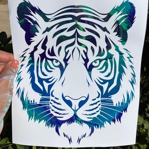 May include: A tiger head decal featuring a white silhouette design layered over a vibrant, iridescent background that shifts between shades of deep blue, teal, and green. The tiger face is detailed with sharp, clean lines and pointed ears.