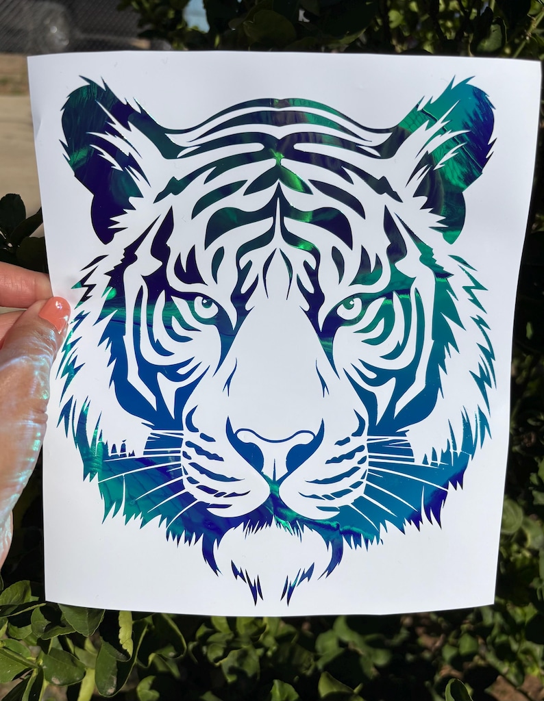 May include: A tiger head decal featuring a white silhouette design layered over a vibrant, iridescent background that shifts between shades of deep blue, teal, and green. The tiger face is detailed with sharp, clean lines and pointed ears.