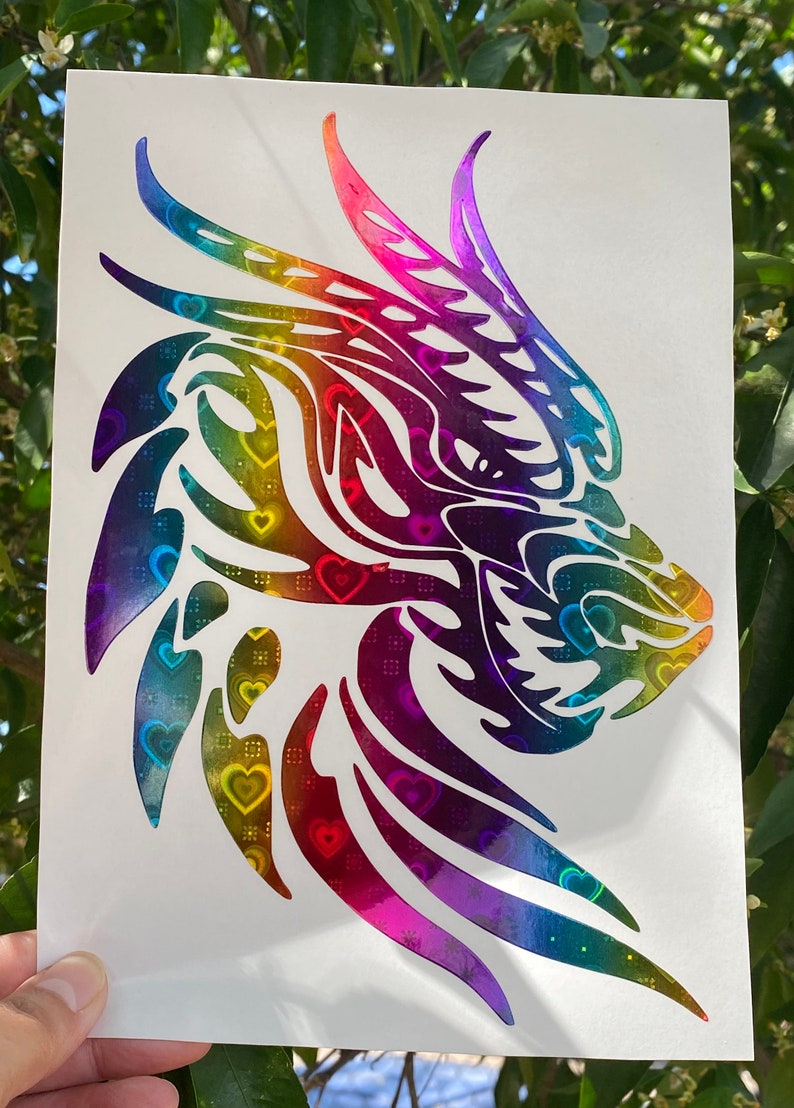 K&ouml;nnte beinhalten: Ein regenbogenfarbener, schimmernder Vinyl-Aufkleber mit einem Drachenkopf vor einem Hintergrund mit Herz-Muster.