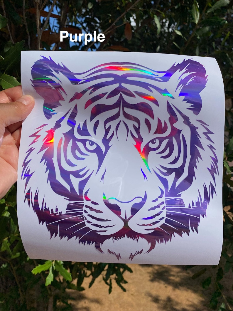 May include: A decal featuring a stylized tiger head graphic with a holographic purple finish on a white backing sheet. The design includes intricate black-toned cutouts for the tiger's facial features and stripes, with the word Purple printed above.
