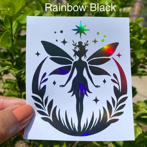 May include: A white sticker with a black fairy design and the text "Rainbow Black". The fairy has iridescent wings and is surrounded by stars and leaves. The sticker is held against a background of green leaves.