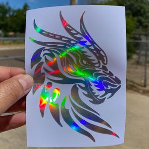 K&ouml;nnte beinhalten: Ein holografischer Aufkleber mit dem Kopf eines Drachen mit einem Tribal-Design. Der Aufkleber befindet sich auf einem wei&szlig;en Hintergrund.
