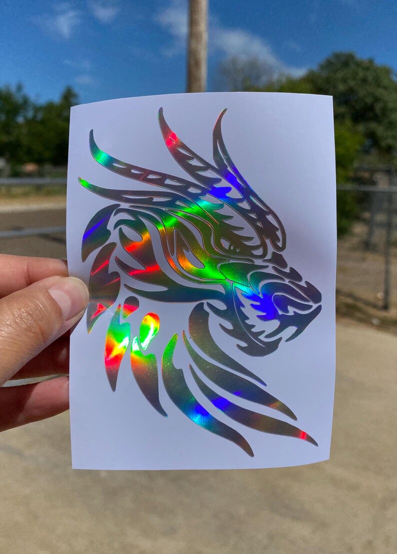 K&ouml;nnte beinhalten: Ein holografischer Aufkleber mit dem Kopf eines Drachen mit einem Tribal-Design. Der Aufkleber befindet sich auf einem wei&szlig;en Hintergrund.