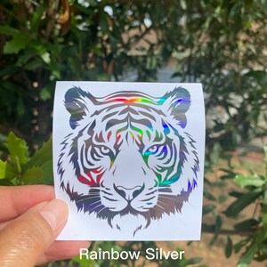 May include: A square white sticker featuring a holographic tiger head graphic with rainbow silver reflective details. The tiger design is rendered in a bold, stylized line art format, showcasing the animal's facial features and stripes.
