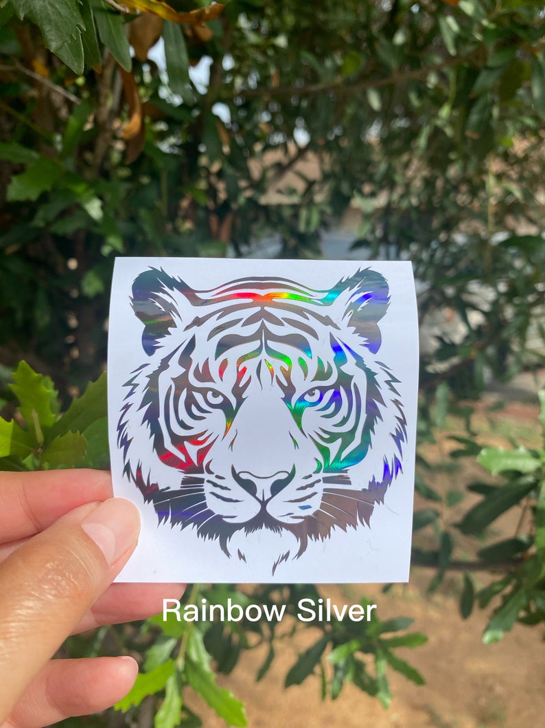 May include: A square white sticker featuring a holographic tiger head graphic with rainbow silver reflective details. The tiger design is rendered in a bold, stylized line art format, showcasing the animal's facial features and stripes.