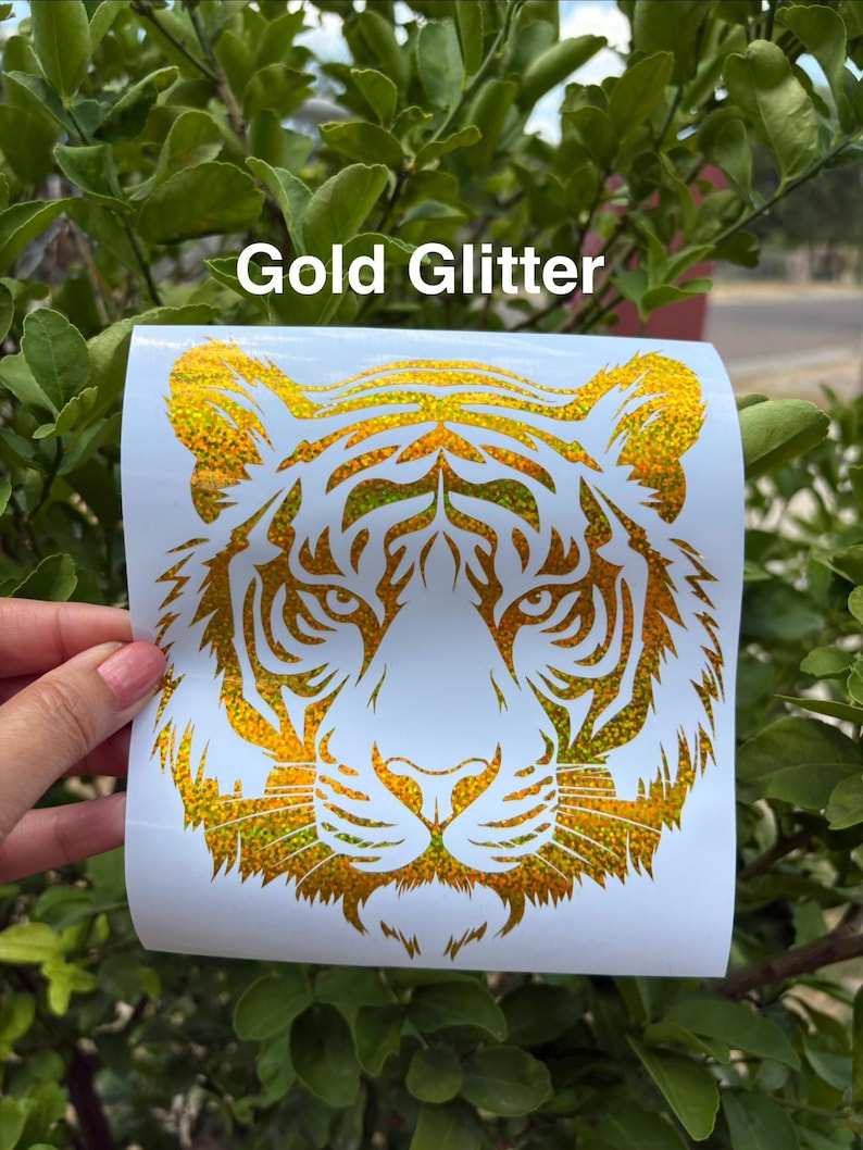 May include: A gold glitter tiger head decal on a white backing sheet with the text Gold Glitter printed above the design. The tiger graphic features intricate cut-out details forming the face, eyes, and whiskers in a shimmering metallic finish.