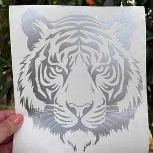 May include: A metallic silver tiger head decal on a white backing sheet. The design features a detailed, symmetrical illustration of a tiger face with sharp, angular lines and distinct facial markings. The text Metallic Silver is visible.