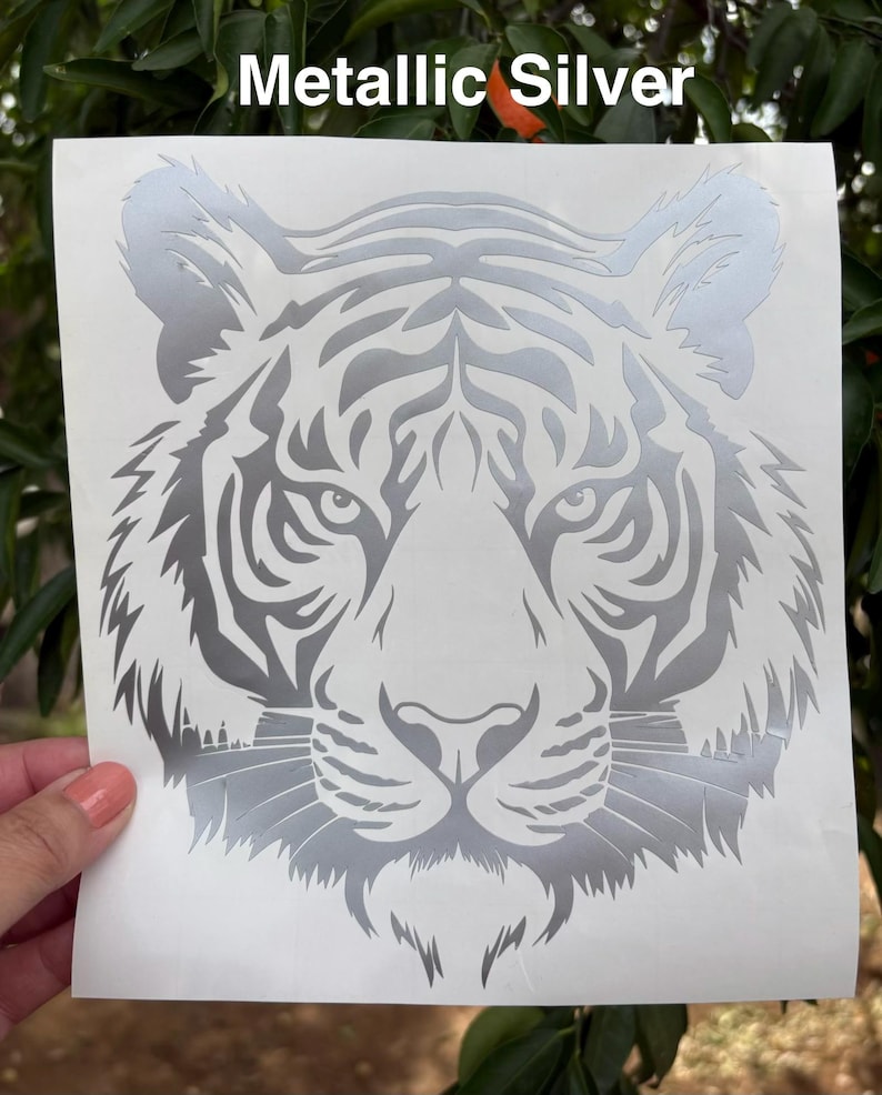 May include: A metallic silver tiger head decal on a white backing sheet. The design features a detailed, symmetrical illustration of a tiger face with sharp, angular lines and distinct facial markings. The text Metallic Silver is visible.