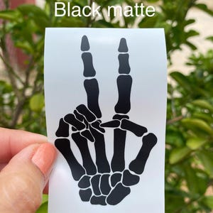 May include: A black matte decal on a white rectangular backing featuring a graphic illustration of a skeleton hand making a peace sign gesture. The text "Black matte" is printed in white above the skeleton hand graphic on the decal sheet.