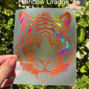 May include: A translucent decal featuring a tiger head graphic with a holographic rainbow orange finish. The tiger design displays intricate black-style line work and facial features. The text Rainbow Orange is printed in white above the tiger.