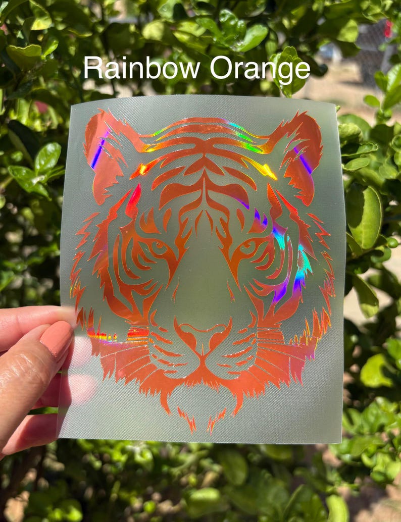 May include: A translucent decal featuring a tiger head graphic with a holographic rainbow orange finish. The tiger design displays intricate black-style line work and facial features. The text Rainbow Orange is printed in white above the tiger.