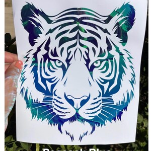 May include: A decal featuring a stylized tiger head design with intricate cutouts. The tiger graphic displays a vibrant, iridescent peacock blue color with a metallic, reflective finish that shifts in tone. The text Peacock Blue is printed below.