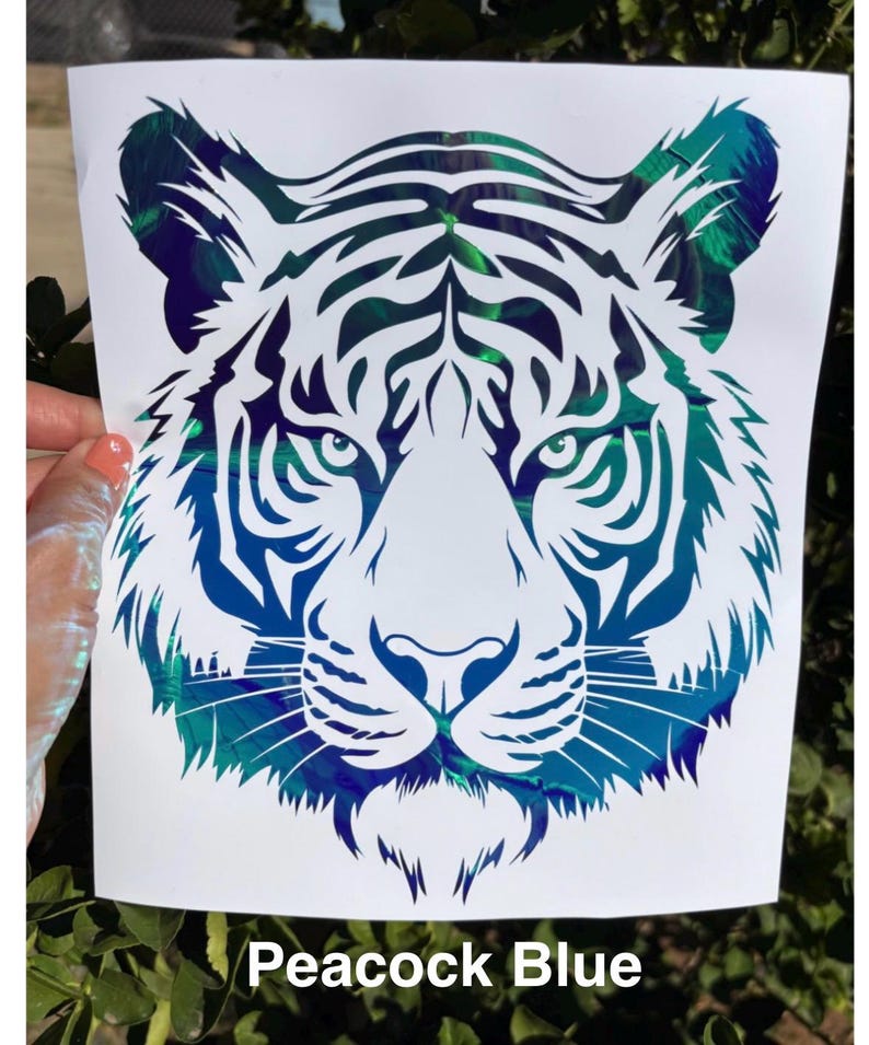 May include: A decal featuring a stylized tiger head design with intricate cutouts. The tiger graphic displays a vibrant, iridescent peacock blue color with a metallic, reflective finish that shifts in tone. The text Peacock Blue is printed below.