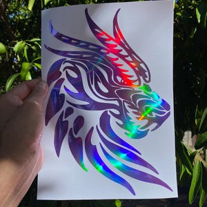 K&ouml;nnte beinhalten: Ein holografischer Vinyl-Aufkleber mit einem Drachenkopf mit Tribal-Design. Der Aufkleber befindet sich auf einem wei&szlig;en Hintergrund.