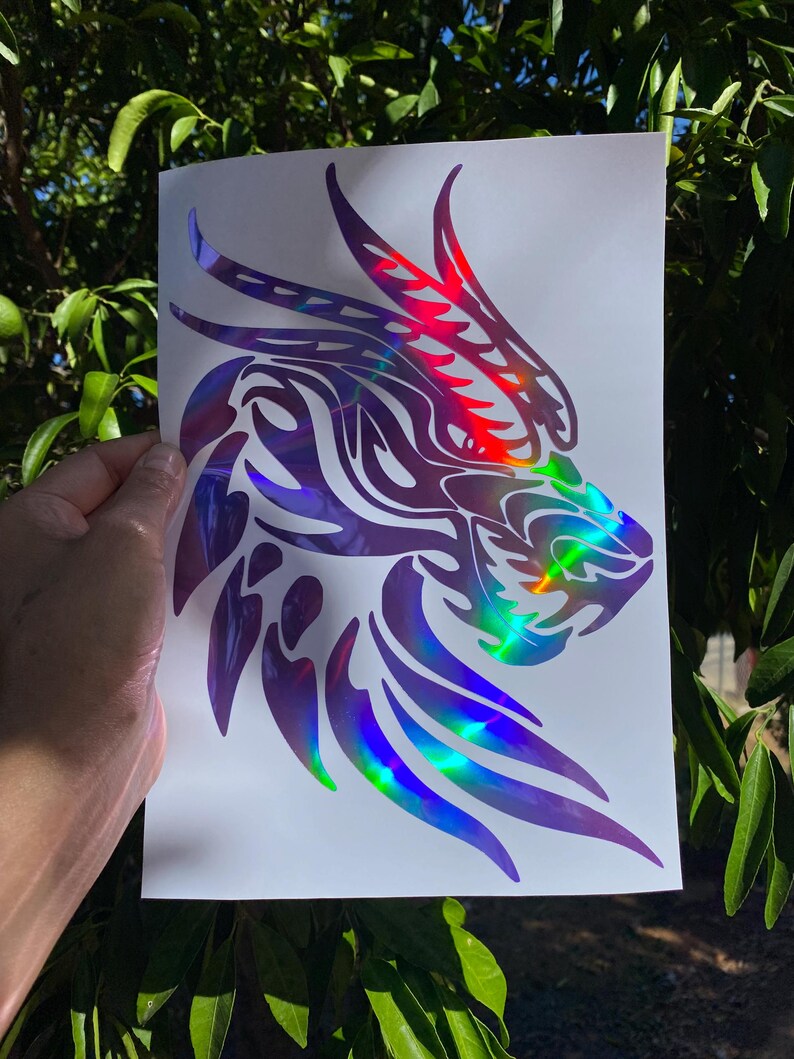 K&ouml;nnte beinhalten: Ein holografischer Vinyl-Aufkleber mit einem Drachenkopf mit Tribal-Design. Der Aufkleber befindet sich auf einem wei&szlig;en Hintergrund.