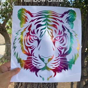 May include: A square white decal featuring a stylized tiger head graphic with a rainbow glitter finish. The design consists of intricate cut-out lines forming the tiger's face, ears, and whiskers. The text "Rainbow Glitter" is printed at the top.