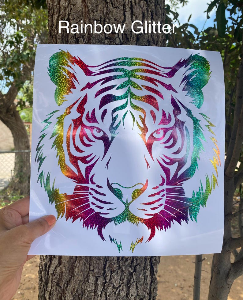 May include: A square white decal featuring a stylized tiger head graphic with a rainbow glitter finish. The design consists of intricate cut-out lines forming the tiger's face, ears, and whiskers. The text "Rainbow Glitter" is printed at the top.